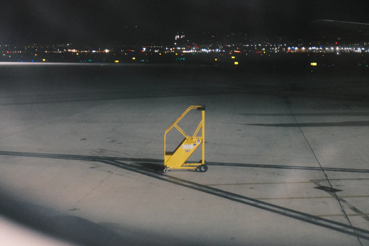 A yellow maintenance ladder standing alone on an empty apron at night, airport lights in the distance