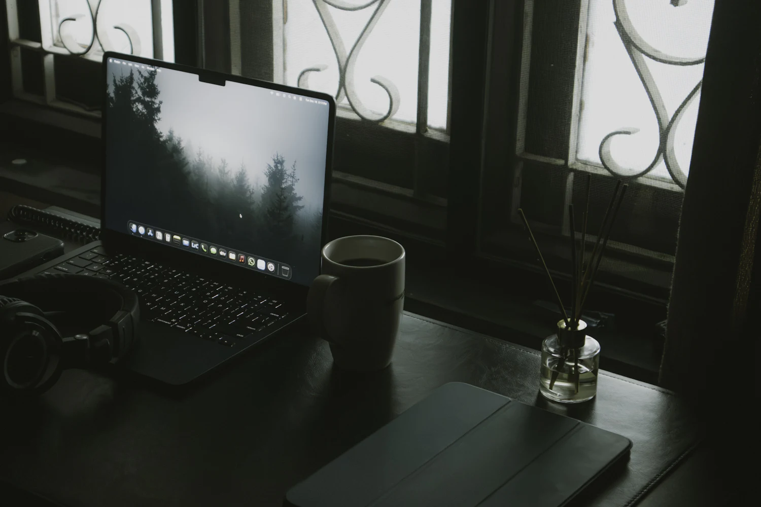 Laptop, headphones and a cup of coffee on a dark desk by a window — a remote workspace at dusk