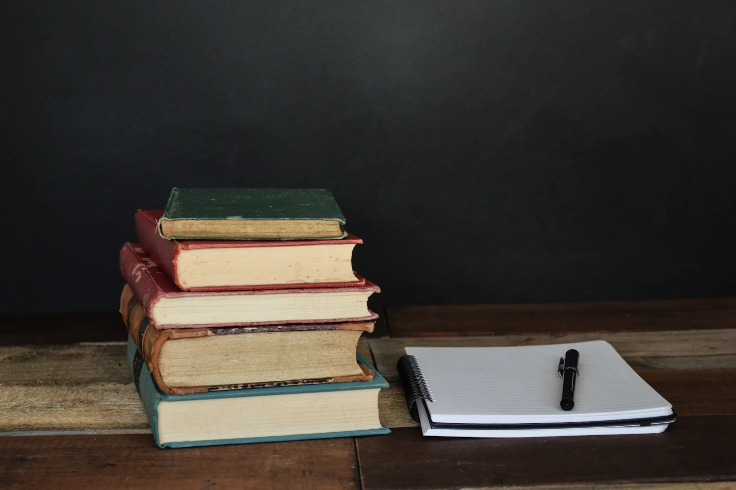 A stack of worn hardback books beside a notepad and pen on a dark wooden desk — the franchising library of Anna Tsirat