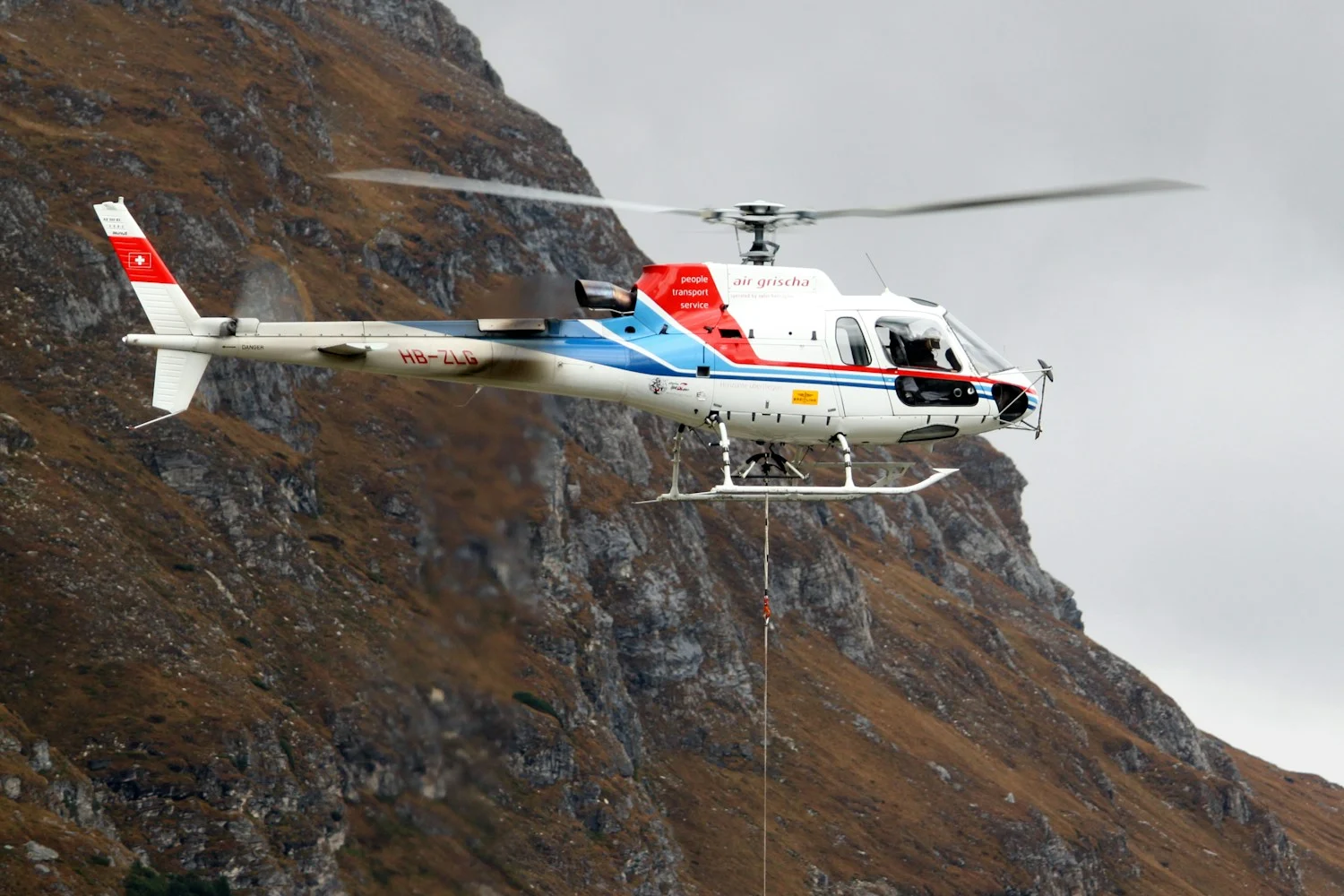A working helicopter with an external load over a mountain slope — illustration for a piece on who counts as a passenger on aerial works