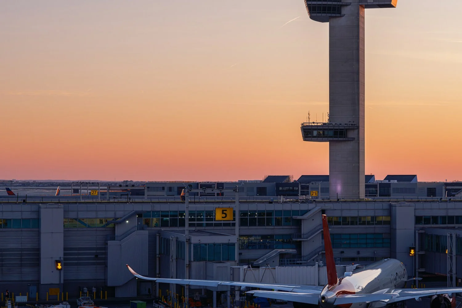 An airport control tower at sunset, with the terminal and an aircraft at the stand below